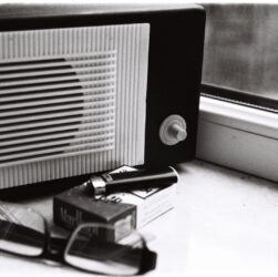 Black and white still life featuring a retro radio, glasses, and cigarette pack by a window.