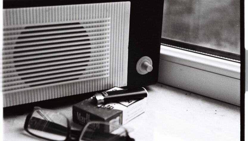 Black and white still life featuring a retro radio, glasses, and cigarette pack by a window.